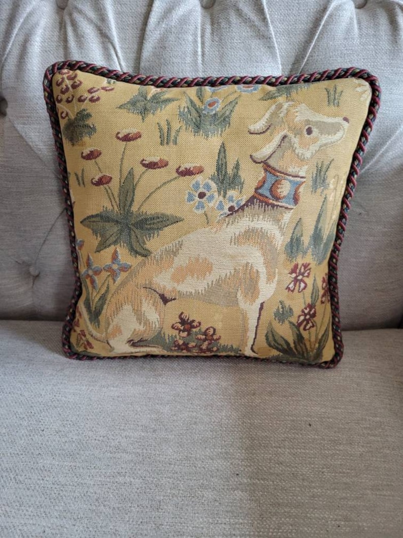 Puede incluir: Una almohada decorativa con un dise&ntilde;o de tapiz que presenta un perro beige con un collar azul y un patr&oacute;n floral sobre un fondo amarillo. La almohada tiene un ribete trenzado marr&oacute;n y rojo.