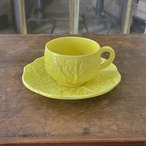 May include: A bright yellow teacup and saucer set. The cup has a handle and sits on a matching saucer. The set is made of ceramic and has a textured, embossed design. The set is on a wooden surface.
