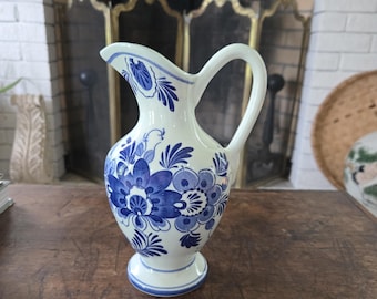 May include: A white ceramic pitcher with a blue floral design. The pitcher has a handle and a spout. The floral pattern includes large, stylized flowers and leaves. The pitcher sits on a wooden surface.