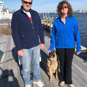 May include: A couple and their dog stand on a wooden dock. The man is wearing a blue jacket, jeans, and sunglasses. The woman is wearing a blue jacket, black pants, and sunglasses. The dog is a German Shepherd with brown fur.