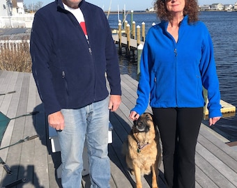 May include: A couple and their dog stand on a wooden dock. The man is wearing a blue jacket, jeans, and sunglasses. The woman is wearing a blue jacket, black pants, and sunglasses. The dog is a German Shepherd with brown fur.