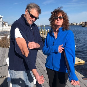 May include: Two people wearing blue fleece jackets with zippers that are open at the elbows. The man is wearing sunglasses and the woman is wearing sunglasses and has curly brown hair.