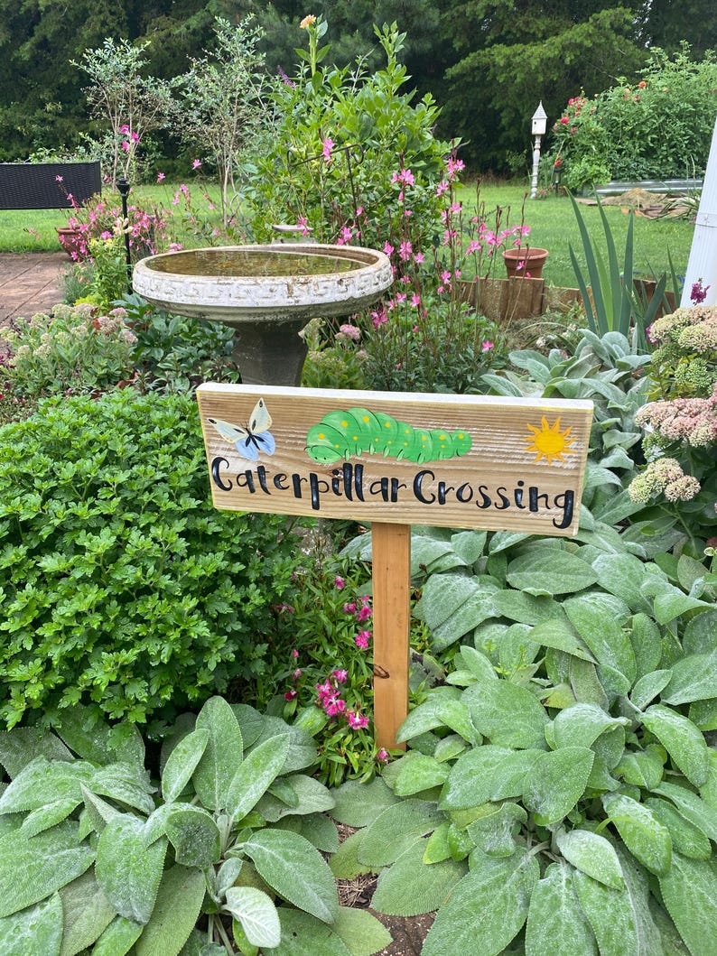 Puede incluir: Un letrero de madera en un jard&iacute;n que dice "Caterpillar Crossing". El letrero presenta una mariposa azul pintada, una oruga verde y un sol amarillo. El letrero est&aacute; montado en una estaca de madera y rodeado de vegetaci&oacute;n y flores.