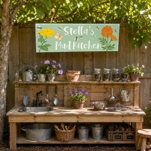 May include: A wooden mud kitchen featuring a light green sign with the text Stella's Mud Kitchen, decorated with painted flowers, bees, and butterflies. The station includes a metal sink, faucet, hanging utensils, pots, and a wicker basket.