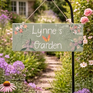 Könnte beinhalten: Ein rechteckiges, salbeigrünes Gartenschild mit dem Text "Lynne's Garden" in weißer Schrift, verziert mit Blumen- und Schmetterlingsillustrationen. Das Schild hängt an einem schwarzen Metallpfosten in einem üppigen Garten.