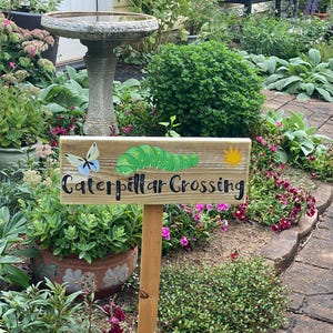 Puede incluir: Un letrero de madera con la inscripci&oacute;n "Caterpillar Crossing" en escritura negra, con una oruga y una mariposa de dibujos animados. El letrero est&aacute; colocado en un jard&iacute;n con un abrevadero y varias plantas.