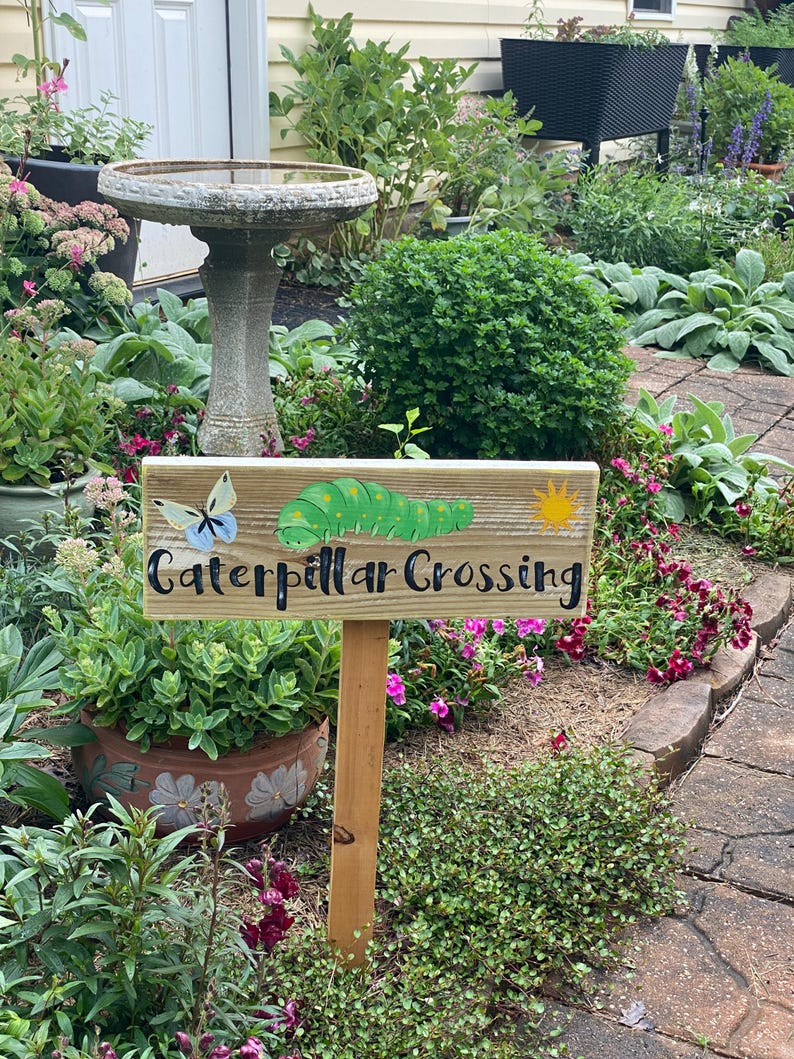 Puede incluir: Un letrero de madera con la inscripci&oacute;n "Caterpillar Crossing" en escritura negra, con una oruga y una mariposa de dibujos animados. El letrero est&aacute; colocado en un jard&iacute;n con un abrevadero y varias plantas.