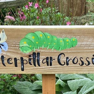 Puede incluir: Cartel de madera con la inscripci&oacute;n "Caterpillar Crossing" en letras negras. Presenta una oruga verde pintada, una mariposa y un sol amarillo. El cartel est&aacute; montado sobre una estaca de madera.