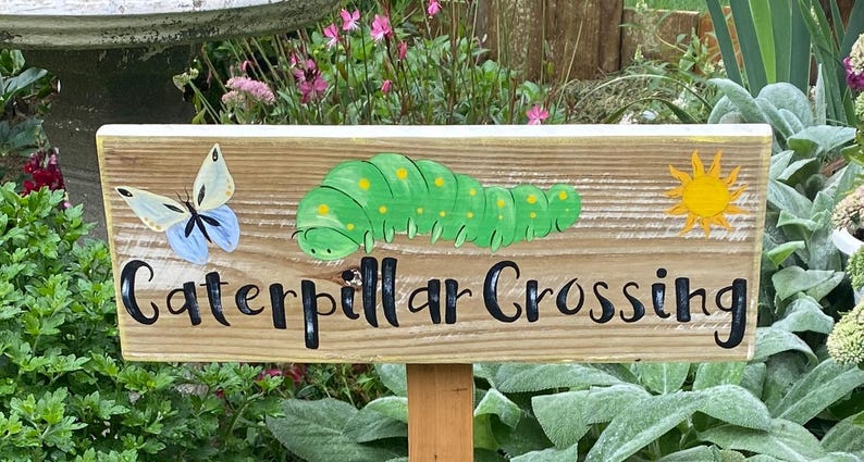 Puede incluir: Cartel de madera con la inscripci&oacute;n "Caterpillar Crossing" en letras negras. Presenta una oruga verde pintada, una mariposa y un sol amarillo. El cartel est&aacute; montado sobre una estaca de madera.