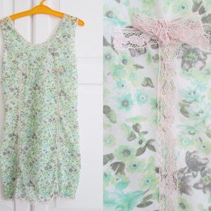 May include: A sleeveless, floral-print slip dress in shades of green, gray, and white, adorned with delicate lace trim and a pink lace bow. The dress is hanging on a yellow hanger against a white background.