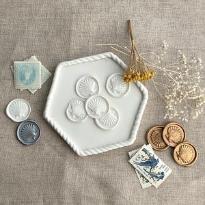 May include: White and gold wax seals with a seashell design. The seals are arranged on a white hexagonal plate with a rope design around the edge. The plate is on a light brown fabric surface.