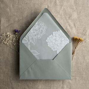 Pu&ograve; includere: Una busta grigia con un disegno floreale bianco sul davanti. Il disegno presenta due fiori di ortensia e foglie.
