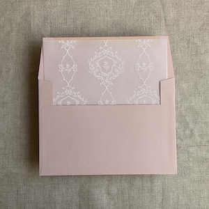 May include: A blush pink envelope with a matching insert featuring a white floral damask pattern. The envelope is open, revealing the patterned insert. The design is elegant and suitable for invitations or announcements.