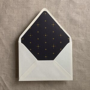 May include: An open envelope with a pointed flap featuring a dark navy blue interior liner. The liner is decorated with a repeating pattern of small, four-pointed gold stars and tiny dots, creating a celestial design against the dark background.