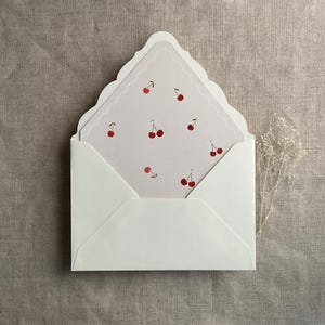 May include: A white envelope with a scalloped edge, containing a pink card with a pattern of red cherries.