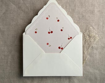 Può includere: Una busta bianca con bordo a festoni, contenente un biglietto rosa con un motivo di ciliegie rosse.