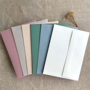 Peut inclure: Un ensemble de six enveloppes de différentes couleurs, dont rose, gris, beige, vert, bleu et blanc. Les enveloppes sont disposées en rangée, l'enveloppe rose à gauche et l'enveloppe blanche à droite.