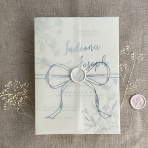 May include: A wedding invitation with a blue ribbon and a wax seal. The invitation is printed on vellum paper and features a floral design. The text on the invitation reads "Indiona & Joseph" and "Saturday, August 8, 2020 at Two Thirty in the Afternoon".