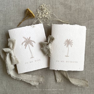 May include: Two off-white greeting cards with a palm tree illustration and the phrases "TO MY WIFE" and "TO MY HUSBAND". The cards are tied with a beige burlap ribbon. Dried flowers and foliage are arranged around the cards.