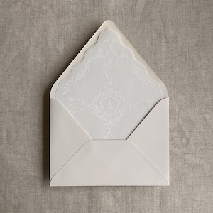 May include: An open cream-colored envelope with a pointed flap, containing a matching cream-colored card. The card features a delicate, white, intricate floral and scrollwork pattern printed across its surface, centered with a shield motif.