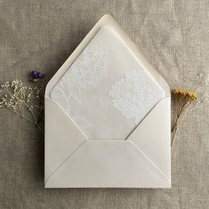 Pu&ograve; includere: Una busta bianca con un motivo floreale bianco sul lembo interno. La busta &egrave; chiusa e si trova su una superficie di tessuto beige.