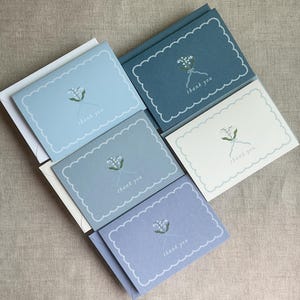 May include: A selection of thank you cards in shades of blue, each with a scalloped edge. The cards display a floral illustration and the words "thank you" in a fine font. White envelopes are included.