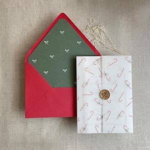 May include: A bright red envelope with a dark green liner featuring a white bow pattern, paired with a translucent vellum wrap decorated with red and white candy canes, secured by a round gold wax seal with a floral embossed design.