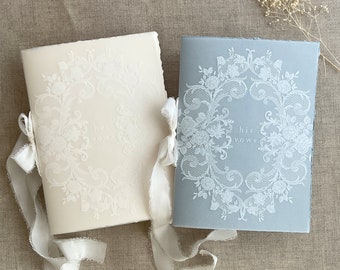 Puede incluir: Dos libros de votos de boda, uno crema y otro azul, con un delicado diseño de encaje. El libro crema dice "her vows" y el libro azul "his vows". Ambos libros están atados con cintas color crema.