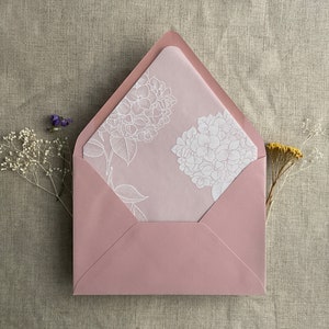 Pu&ograve; includere: Una busta rosa con un disegno floreale bianco sul davanti. La busta &egrave; fatta di carta e ha una linguetta in cima.