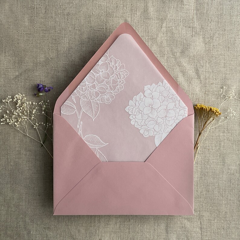 Pu&ograve; includere: Una busta rosa con un disegno floreale bianco sul davanti. La busta &egrave; fatta di carta e ha una linguetta in cima.