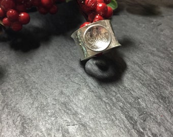 May include: A silver ring with a textured, circular design. The ring is wide and has a hammered finish.