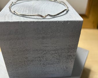 May include: A silver bangle bracelet with a wavy, organic design. The bracelet is displayed on a gray textured block.