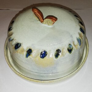 Könnte beinhalten: Eine weiße Keramik-Butterdose mit Deckel. Der Deckel hat ein dekoratives Schmetterlingsdesign in Braun und Gold. Die Dose hat eine weiße Glasur mit blauen und braunen Akzenten.