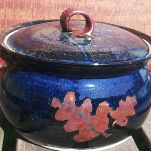 Puede incluir: Una cacerola de cerámica azul con tapa y dos asas. La cacerola tiene un diseño de hoja de roble roja y naranja.