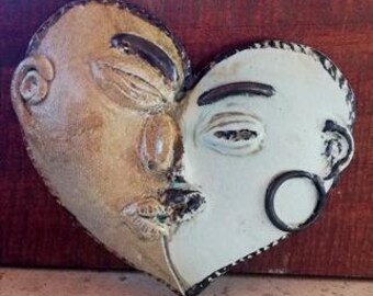 May include: A heart-shaped ceramic wall hanging featuring two faces, one tan and the other white. The tan face has closed eyes and a pursed mouth, while the white face has one open eye and a circular earring. The edges are outlined in black.