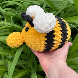 May include: A handmade, plush bee toy with yellow and black stripes, white wings, and black antennae. The bee is made of soft yarn and has black button eyes. The toy is held in a person's hand against a background of green foliage.