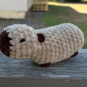 May include: A plush, handmade capybara toy. The body is a light beige color, with a darker brown head, ears, and feet. The capybara has a small black eye and is made of a soft, textured material.