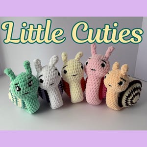 May include: Five crocheted snail toys in various colors: green, white, yellow, pink, and tan. Each snail has a smiling face and a spiral shell. The text "Little Cuties" is above the snails.