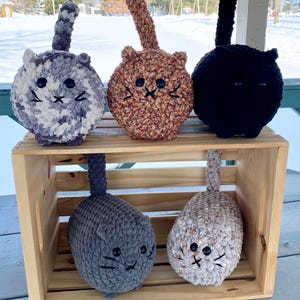 May include: Five crocheted cat plushies in a wooden crate. The cats are gray and white, brown and white, black, gray, and beige. They have black eyes and embroidered whiskers.