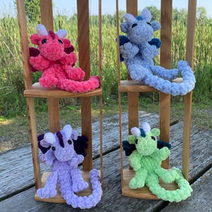 May include: Four handmade crochet dragons in pink, blue, purple, and green, each with unique wing and horn colors. The plush toys are displayed on small wooden chairs, with a natural outdoor background.