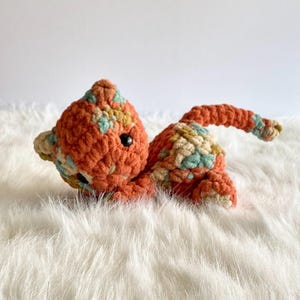 May include: A crocheted cat toy with a multi-colored pattern in shades of orange, green, and beige. The cat is lying on its belly with its tail curled up.