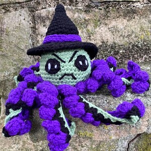 May include: A handmade crochet octopus doll wearing a black witch hat with a purple band. The octopus has a green head with black eyes and a frowning mouth. Its tentacles are purple with black accents.