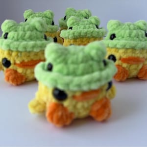 May include: A group of five handmade crochet ducklings, each wearing a green frog hat. The ducklings are yellow, orange, and green, with black eyes. The crochet toys are arranged on a white surface.