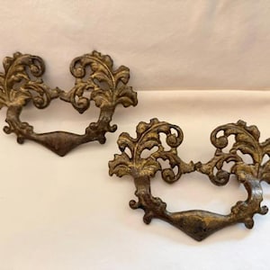 May include: Two antique bronze-colored drawer pulls with ornate, scrolling leaf designs. The pulls have a curved handle and decorative flourishes, perfect for adding a vintage touch to furniture. Each pull measures approximately 6 inches wide.