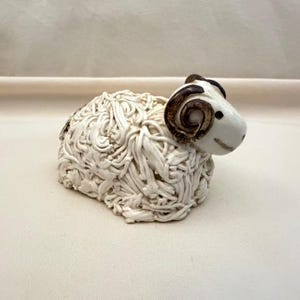 May include: A ceramic ram figurine with a white body and a detailed, textured wool-like appearance. The ram's head is white with brown horns and a smiling expression. The figurine is a decorative item.