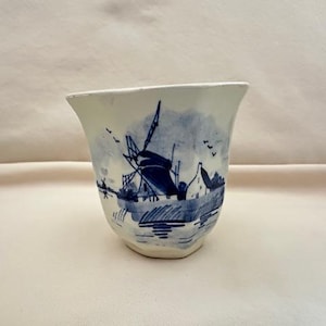 May include: A white ceramic cup with a flared rim, adorned with a blue and white windmill scene. The design includes a windmill, buildings, and water, creating a classic, nautical aesthetic. The cup has a slightly irregular shape.
