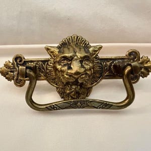 May include: Antique brass drawer pull featuring a detailed lion's head in the center. The pull has a decorative bar with leaf accents and a curved handle. The hardware is a warm, aged brass color.