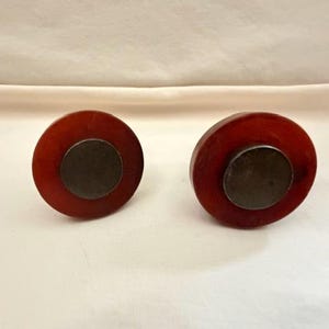 May include: Two circular drawer knobs featuring a deep reddish-brown translucent outer ring and a flat, dark metallic center disc. The design consists of a smooth, polished finish on the outer edge with a contrasting matte dark metal inlay.