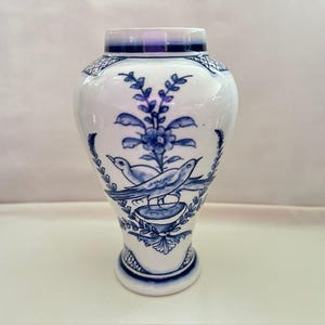 May include: A white ceramic vase with blue floral and bird designs. The vase has a rounded body, a flared base, and a narrow neck with a purple rim. The blue designs include flowers, leaves, and two birds perched on a decorative element.