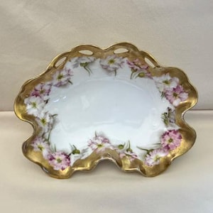 May include: An oval-shaped, white porcelain dish with a scalloped, gold-painted rim. The dish is decorated with pink and white floral patterns. The rim has small, decorative cut-out holes. A delicate and elegant piece.